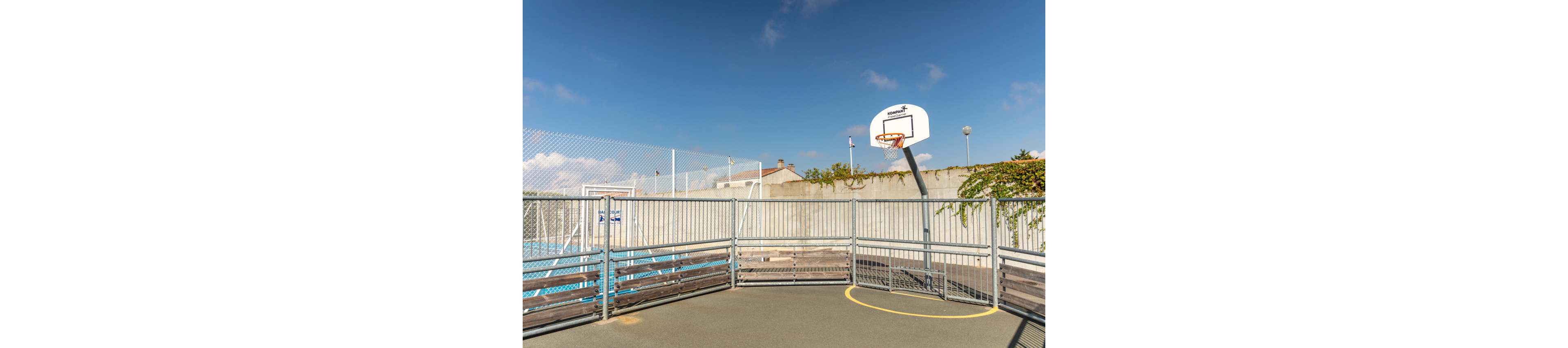 An empty outdoor basketball court features a single hoop mounted above a high concrete wall. Metal railings and fencing enclose the playing area, creating a compact urban sports space. A clear blue sky with scattered clouds provides a bright, sunny backdrop. The scene feels quiet and ready for recreational play.