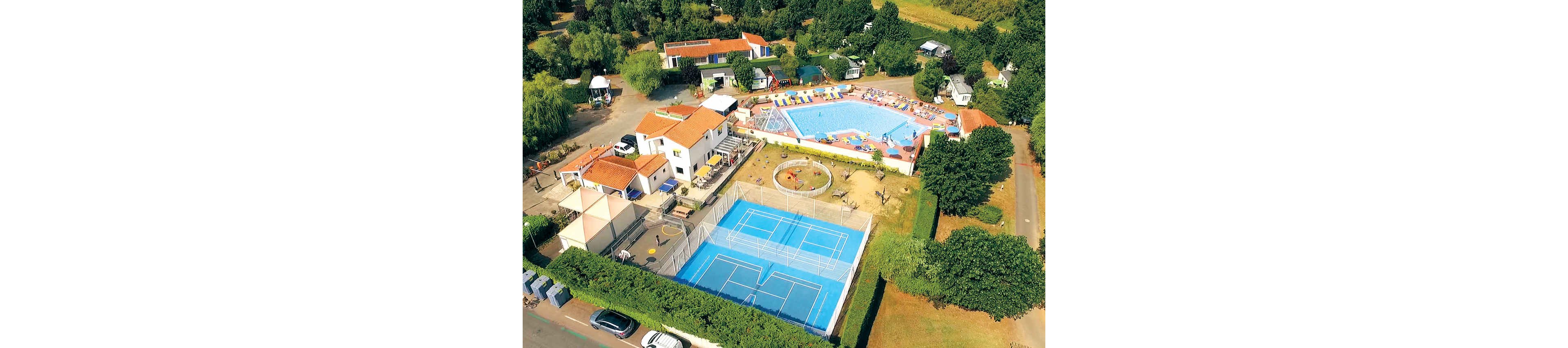 A large holiday resort is captured from above, showing a bright blue swimming pool surrounded by colorful sun loungers. Next to the pool is a fenced multi-court sports area with tennis or basketball markings. A small circular playground and several white-roofed buildings with orange tiles are visible around the central facilities. The complex is bordered by trees, greenery, and parked cars, giving a relaxed vacation atmosphere.