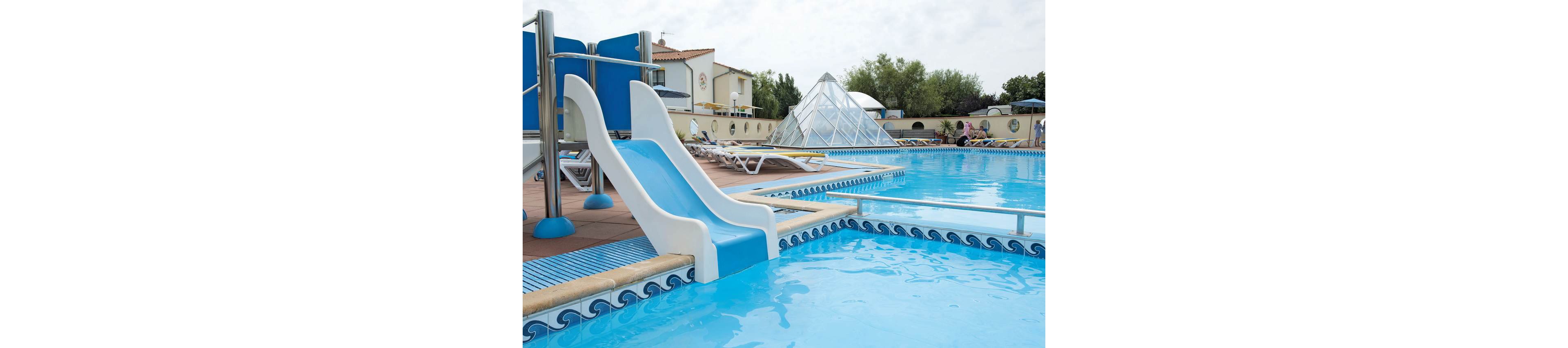 A young boy in red swim trunks slides down a white waterslide into a bright blue outdoor swimming pool. The scene appears to be at a vacation resort with buildings, trees, and a glass pyramid structure in the background. A yellow inflatable ring floats in the foreground near the pool edge. The atmosphere is relaxed and playful, suggesting a warm day of family leisure by the water.