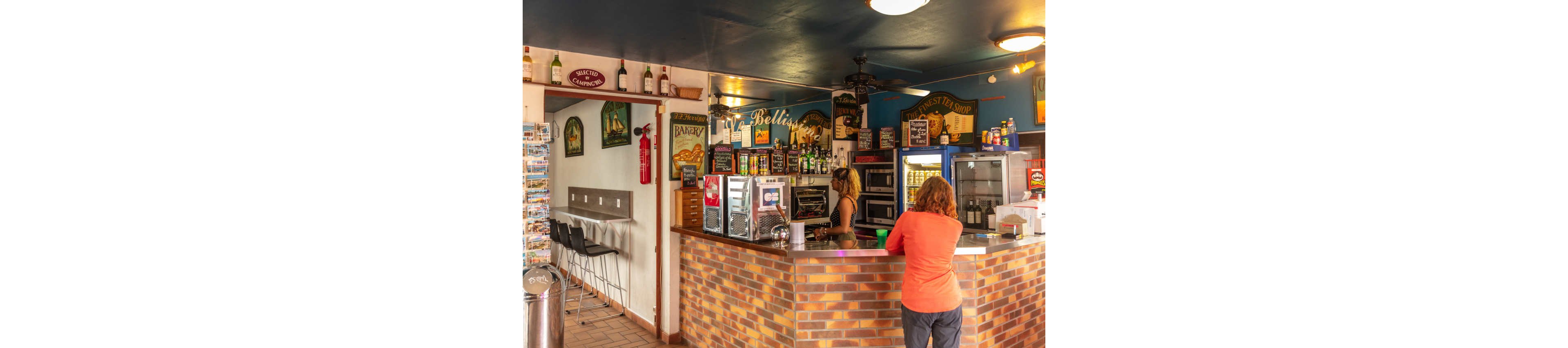 An indoor café bar features a brick-front counter with various drink coolers and shelves of bottles behind it. A woman stands behind the counter serving a customer who is leaning on the bar, while another seating area with barstools is visible to the side. The walls are decorated with colorful signs, menus, and beverage advertisements, creating a casual, welcoming atmosphere. Warm lighting and mixed wood and metal furnishings emphasize the relaxed, social setting.