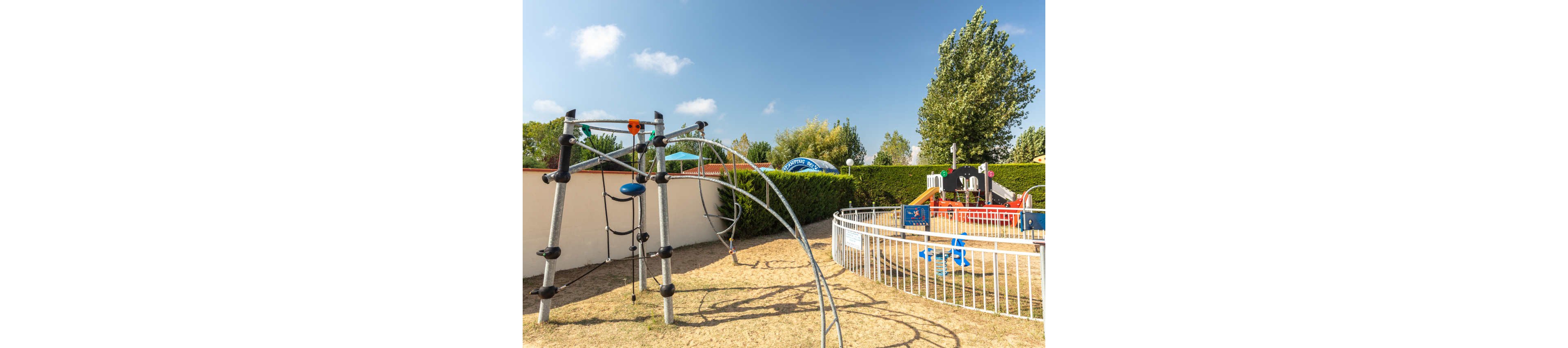 An outdoor playground features a modern metal climbing frame on a dry, sandy surface. A small fenced play area with colorful equipment and a blue spring rider sits nearby. Green hedges and trees surround the playground, with a clear blue sky and scattered clouds overhead. The scene suggests a bright, sunny day in a family-friendly recreational area.