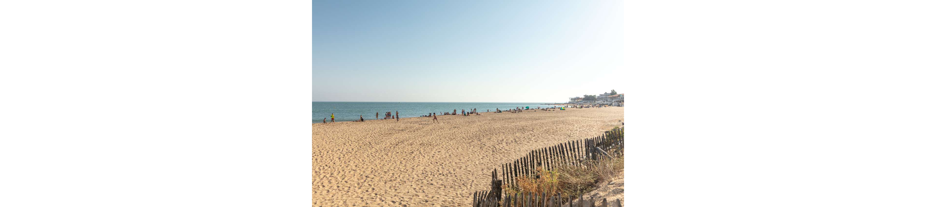 A wide sandy beach stretches along a calm sea under a clear blue sky. Small groups of people relax and walk near the shoreline, enjoying the warm weather. The foreground features a rustic wooden fence and dry dune vegetation leading down to the open sand. The horizon is bright and hazy, giving the scene a peaceful, summery atmosphere.