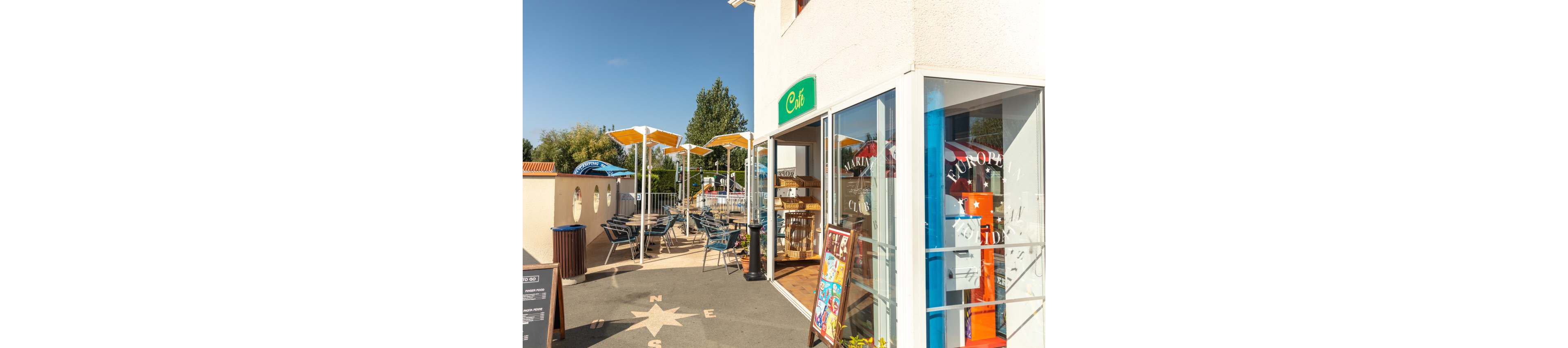 A small café with glass walls opens onto a sunny outdoor terrace. Several empty tables and chairs are arranged under yellow shade structures, suggesting a relaxed dining area. A standing menu board and various colorful signs are visible near the entrance. The scene is bright and inviting, with clear blue skies and trees in the background.