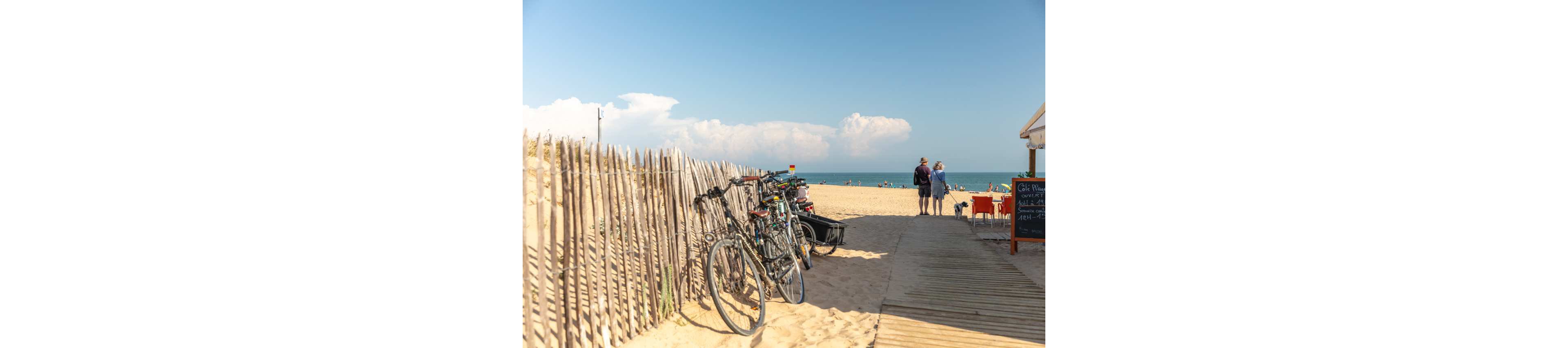 Several bicycles are parked against a wooden fence beside a sandy beach. A wooden boardwalk leads toward the shoreline where two adults stand looking at the sea. Beachgoers are visible in the distance near the water under a bright blue sky with scattered clouds. Warm sunlight creates a relaxed summer atmosphere along the coast.