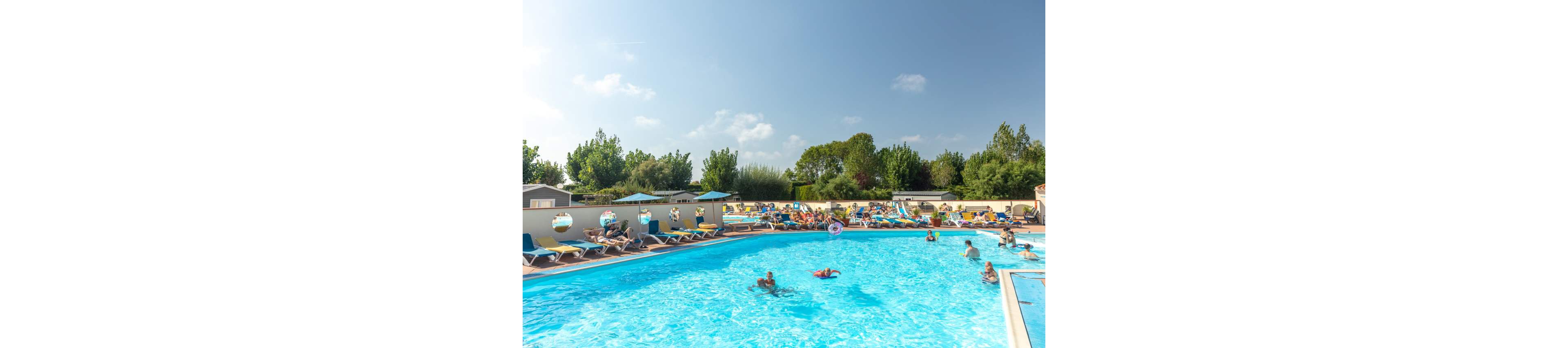 An outdoor swimming pool is filled with people enjoying a sunny day. Adults and children relax on sun loungers and play in the bright blue water. Colorful inflatables and umbrellas add to the lively holiday atmosphere. Trees and low buildings surround the pool area under a clear blue sky.