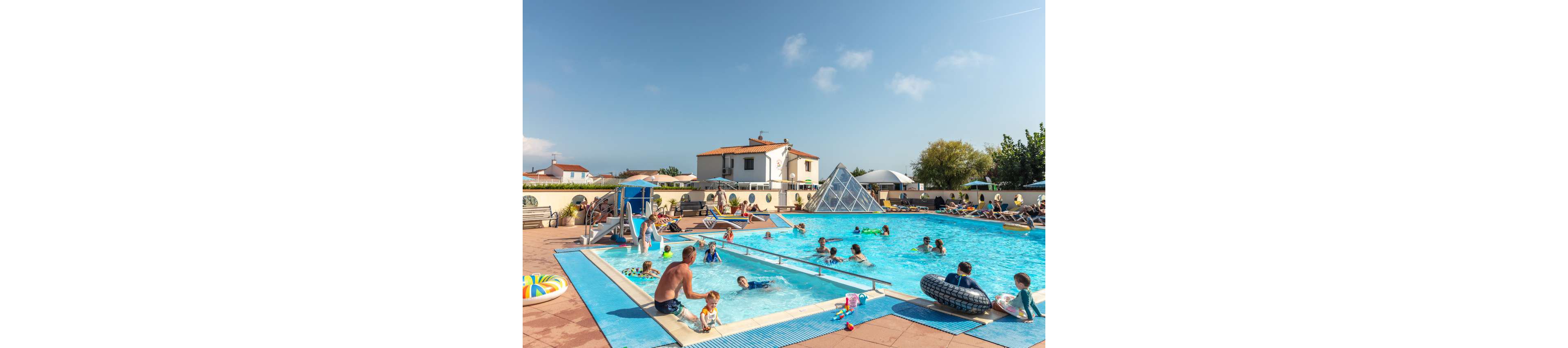 A busy outdoor swimming pool is filled with families and children playing in the water. Adults relax on sun loungers around the pool while kids splash, swim, and use inflatable toys. The scene is set in a residential or holiday complex with low white buildings and a glass pyramid structure in the background. Bright daylight and clear skies create a warm, summery vacation atmosphere.