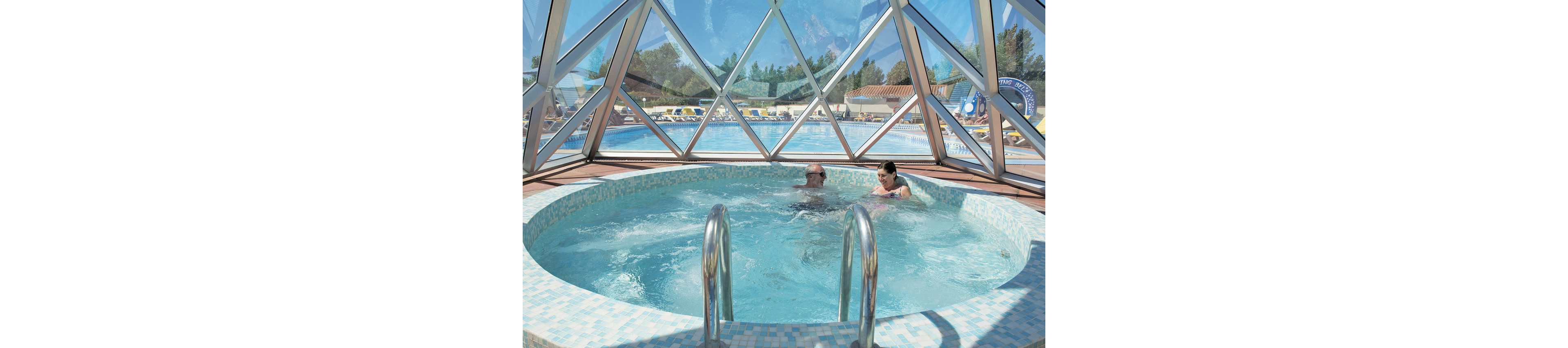 Two people relax in a circular hot tub enclosed by a glass dome structure. The bubbling water suggests a warm, spa-like experience. Outside the dome, a large swimming pool, sun loungers, and umbrellas indicate a resort or holiday setting. The scene is bright and sunny, creating a leisurely vacation atmosphere.