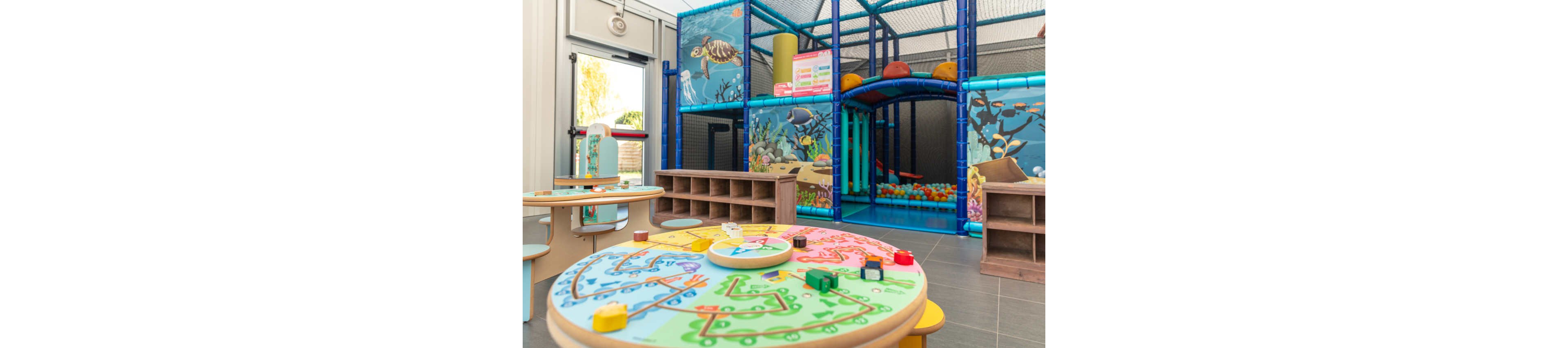 A bright indoor playroom features round activity tables with colorful board games and wooden pieces in the foreground. In the background, a large soft-play climbing structure and ball pit are enclosed with netting. The walls and play area are decorated with an underwater ocean theme, including fish and sea plants. Natural light enters from a nearby window, creating a cheerful and inviting atmosphere for children.
