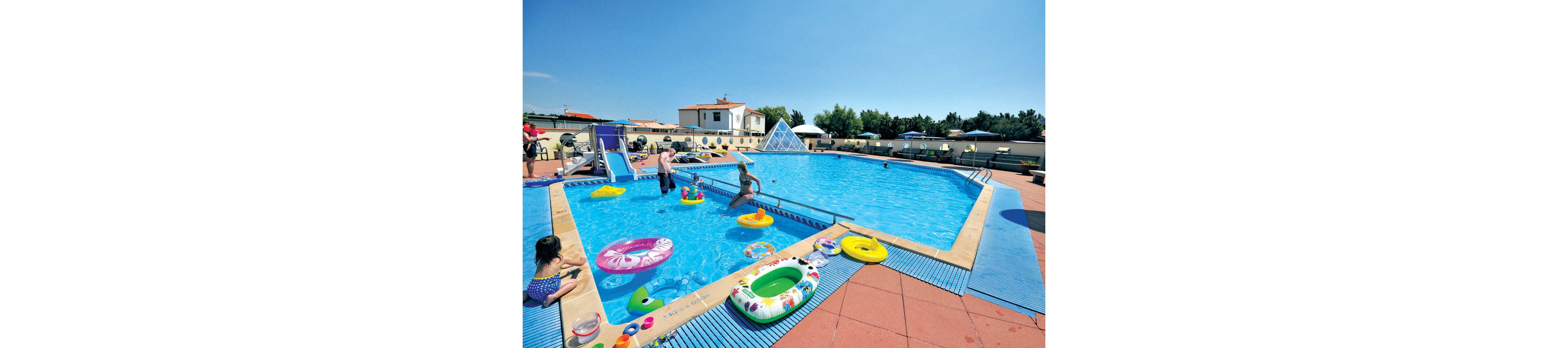An outdoor swimming pool is filled with families and children playing in the water. Colorful inflatable rings and pool toys float on the bright blue surface. A small slide leads into the shallow end while adults supervise from the poolside. The scene is set under a clear blue sky with surrounding buildings and lounge chairs in the background, creating a relaxed vacation atmosphere.