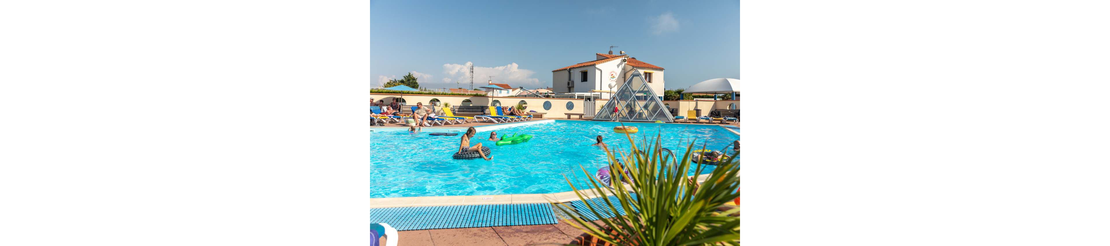 Families and children enjoy a bright day in a large outdoor swimming pool. People float on inflatable rings and loungers while others sit on deck chairs around the pool. A white building and a glass pyramid structure rise in the background under a clear blue sky. Green plants in the foreground add a tropical holiday atmosphere to the scene.