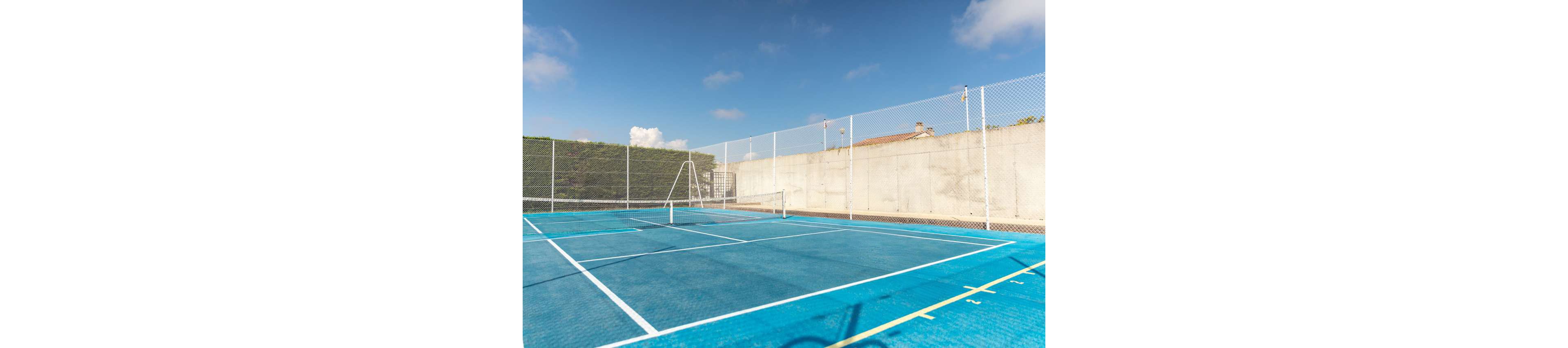 Two adults play a game of tennis on a bright blue outdoor court. The woman in the foreground prepares to hit the ball while facing her opponent across the net. The court is surrounded by high chain-link fencing and tall hedges, with a concrete wall and buildings in the background. Clear blue sky and sunlight create a bright, active atmosphere.