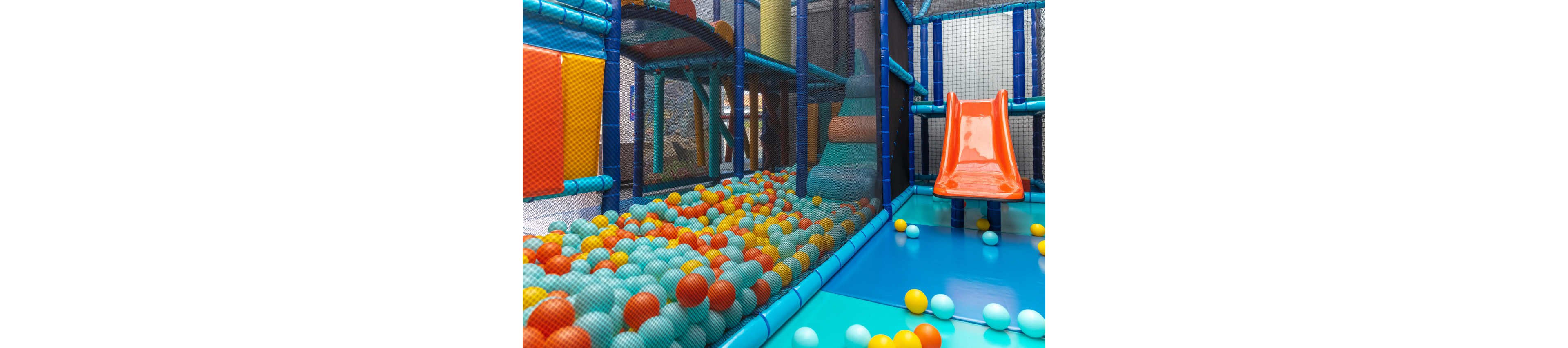 A bright indoor playground features a large ball pit filled with multicolored plastic balls. A shiny orange slide leads down into the pit, surrounded by padded blue flooring and safety netting. Soft play structures and climbing areas are visible in the background, creating a fun and secure environment. The scene suggests a children’s play area in an indoor amusement center or family entertainment venue.