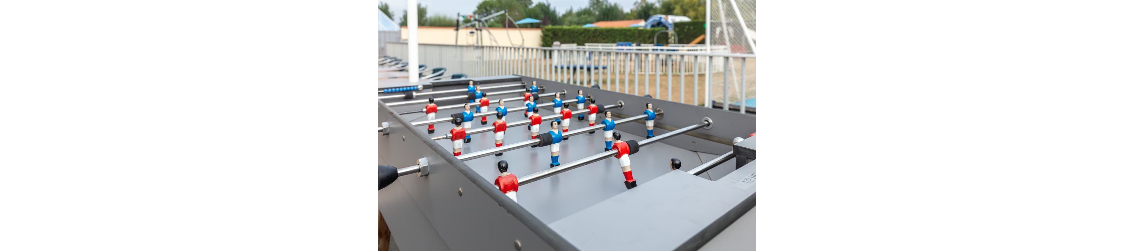 A gray foosball table with red and blue player figures is set up outdoors. The table is positioned near a fenced area with a playground and sports equipment visible in the background. The scene suggests a leisure or recreation area, possibly at a park or holiday resort. Soft daylight and muted colors create a relaxed, casual atmosphere.