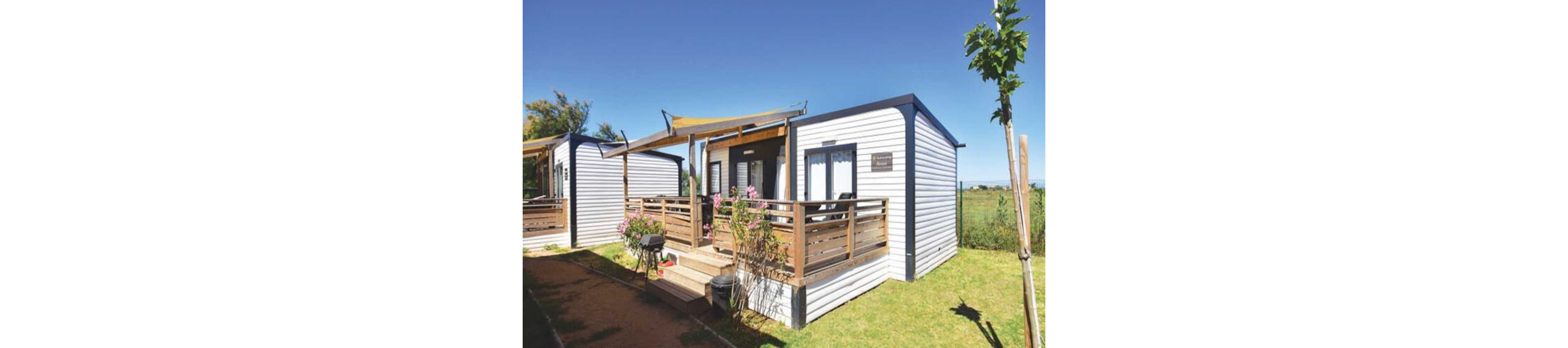 A compact modern mobile home with white siding and dark trim stands in a sunny outdoor setting. The unit features a raised wooden deck with railing, potted plants, and a small grill near the steps. A fabric shade canopy stretches above the terrace, providing partial cover. The surrounding area includes green grass, a dirt path, and open landscape under a clear blue sky.