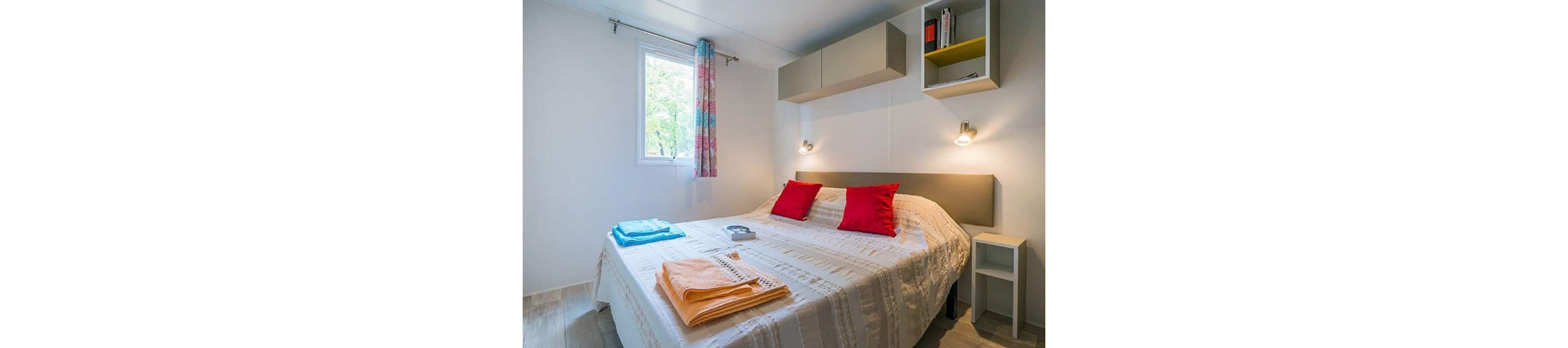A neatly made double bed with a textured white bedspread is the focal point of this compact bedroom. The room features colorful accents, including two red pillows and folded orange and blue towels placed on the bed. A small window with patterned curtains lets in natural light, illuminating the light wood floor and white walls. Wall-mounted reading lights, overhead storage cabinets, and a small bedside table complete the modern, minimalist decor.