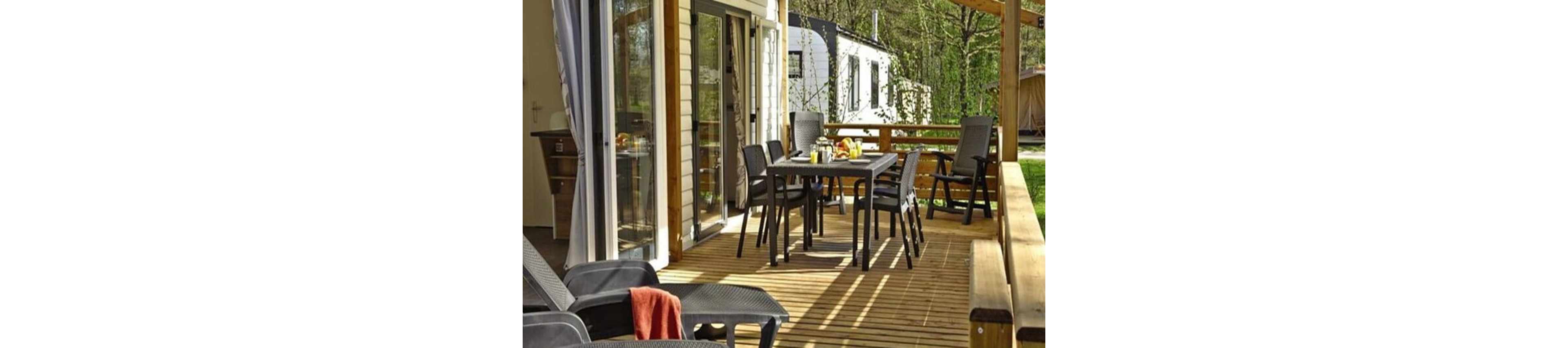 A spacious wooden terrace is furnished with a dark outdoor dining table and several chairs. Sunlight casts strong shadows across the deck, creating a warm and inviting atmosphere. Sliding glass doors open from the house onto the terrace, where breakfast items are set on the table. In the background, a caravan and trees suggest a holiday park or campsite setting.