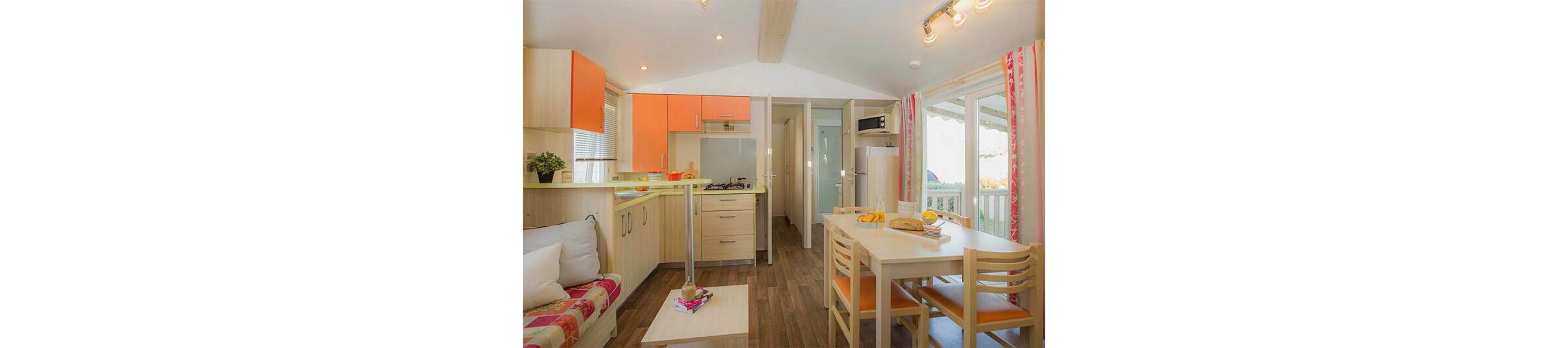 Compact open-plan kitchen and dining area inside a modern holiday cabin. Light wood cabinetry with orange accents frames a small stove, sink, and refrigerator. A simple dining table with chairs sits beside large windows and a sliding door leading to a balcony. A cozy sofa with colorful cushions and a coffee table complete the inviting interior. Warm lighting and natural daylight create a cheerful, welcoming atmosphere.