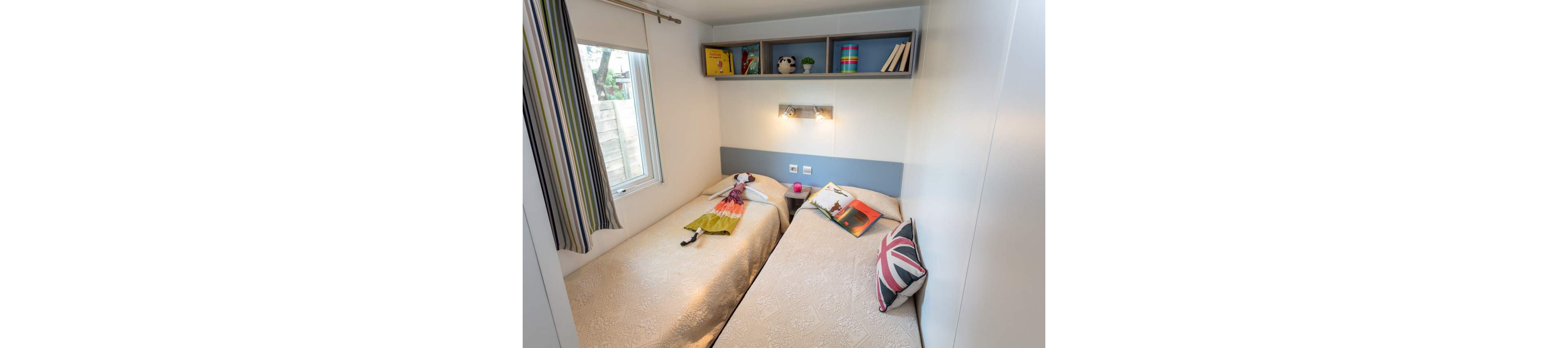 Compact modern bedroom featuring two single beds with light patterned bedspreads. A window with striped curtains brings natural light into the small space. Above the beds, open shelves display books, toys, and decorative items, adding bright color accents. A Union Jack pillow and a laid-out dress suggest a lived-in, welcoming atmosphere.