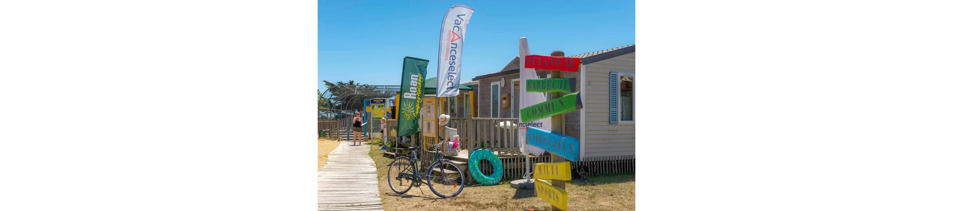 A sunny beachside holiday campsite features wooden cabins and a wooden boardwalk. Colorful directional signs and tall flags with visible text such as "vacanceselect" and "nani" add to the lively atmosphere. A bicycle is parked near the path, and a green inflatable ring rests against the railing. People in swimwear walk toward the beach under a clear blue sky, suggesting a relaxed summer vacation setting.