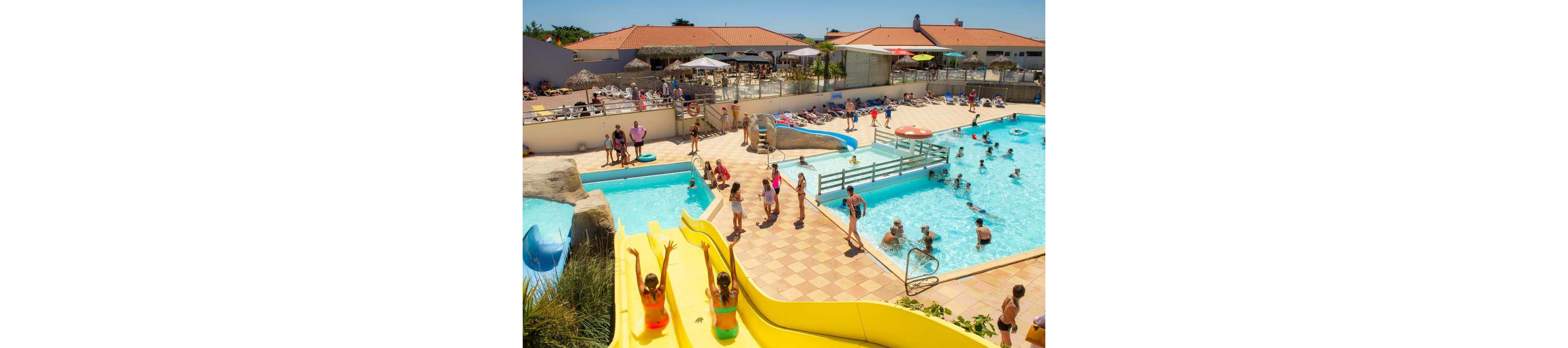 An outdoor water park is filled with families enjoying multiple swimming pools and slides on a sunny day. Children play on bright yellow water slides while others splash in the clear blue pools. Adults relax on poolside loungers and walk along tiled pathways surrounding the aquatic area. The scene suggests a lively vacation resort atmosphere with warm weather and clear skies.