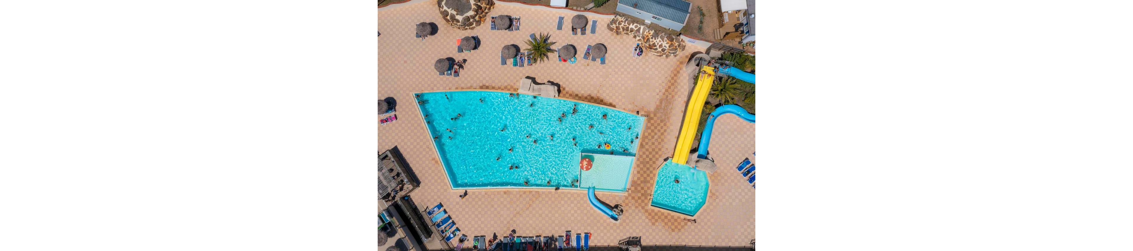 A large outdoor swimming pool complex is seen from above on a sunny day. Numerous people are swimming and playing in the main pool and smaller adjoining pool. Bright yellow and blue water slides curve into the water, adding a sense of fun and motion. Surrounding the pools are sun loungers, umbrellas, and palm trees on a tiled deck, creating a relaxed vacation atmosphere.