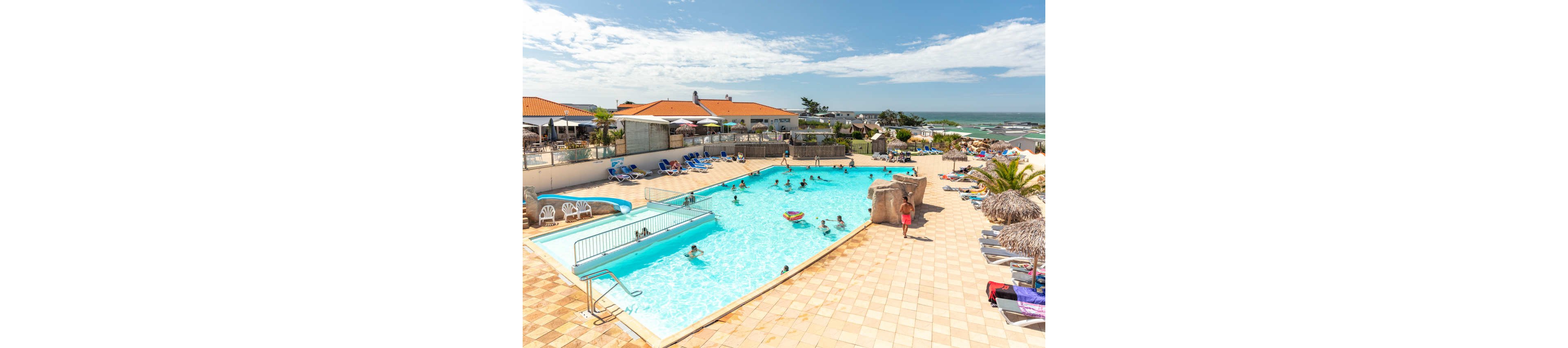 An outdoor swimming pool at a seaside resort is filled with people enjoying the water under bright sunshine. Sun loungers and umbrellas line the tiled pool deck, with guests relaxing and sunbathing. Low-rise buildings with red-tiled roofs surround the pool area, and the ocean is visible in the background beyond a sandy shoreline. The scene conveys a relaxed vacation atmosphere with clear blue skies and calm waves.