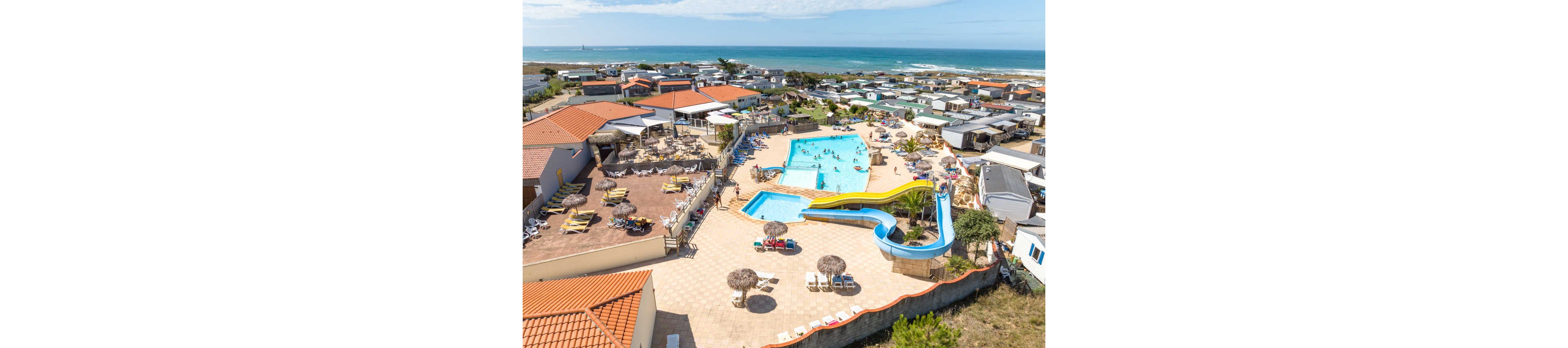 A large coastal campsite features a central outdoor swimming pool complex with bright yellow and blue water slides. Guests relax on sun loungers and under thatched umbrellas around the pool terrace. Rows of mobile homes and caravans extend toward the shoreline, with the ocean visible in the background. The scene suggests a sunny summer holiday atmosphere with families enjoying leisure time by the sea.
