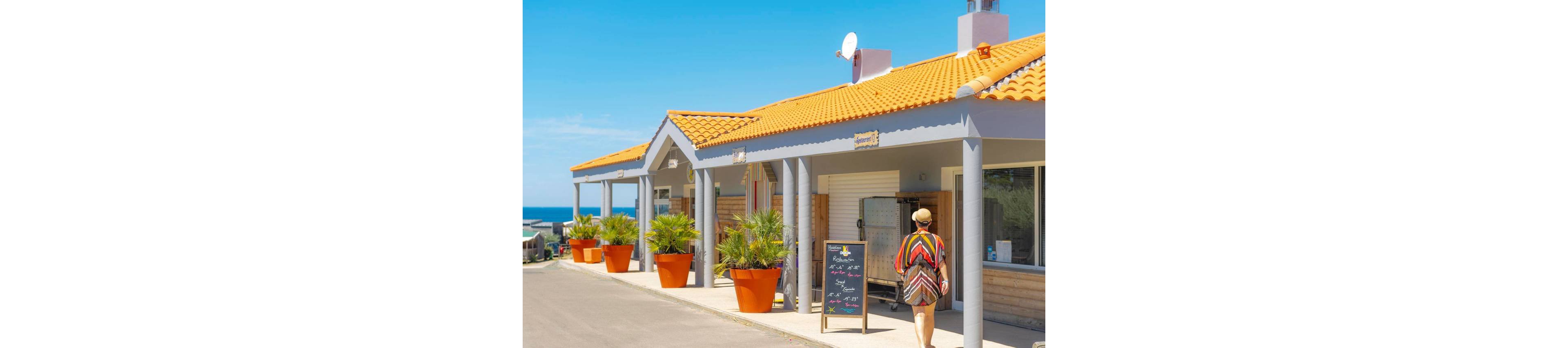 A woman in a patterned dress and hat walks along a sunny promenade past a small seaside cafe. The building features bright orange roof tiles, white pillars, and large orange planters with palm plants. A chalkboard menu stands outside the entrance, and the blue ocean is visible in the background under a clear sky. The scene evokes a relaxed holiday atmosphere in a coastal town.