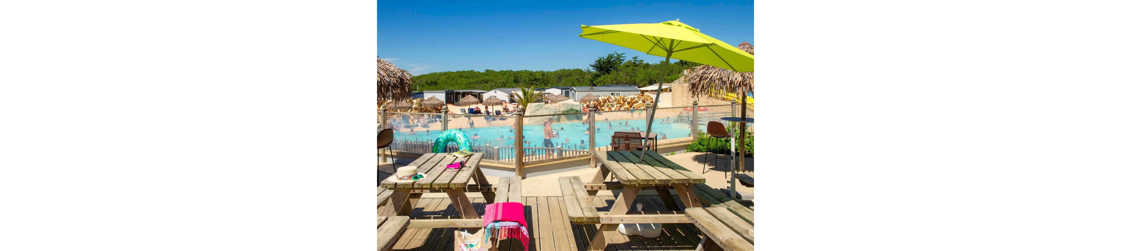 An outdoor swimming pool at a campsite is viewed from a shaded wooden deck with picnic tables in the foreground. Families and children enjoy the bright blue pool surrounded by a glass safety fence and tropical-style umbrellas. Colorful towels, bags, and pool toys add vibrant accents to the sunlit scene. Mobile homes and lush greenery frame the background under a clear blue sky.