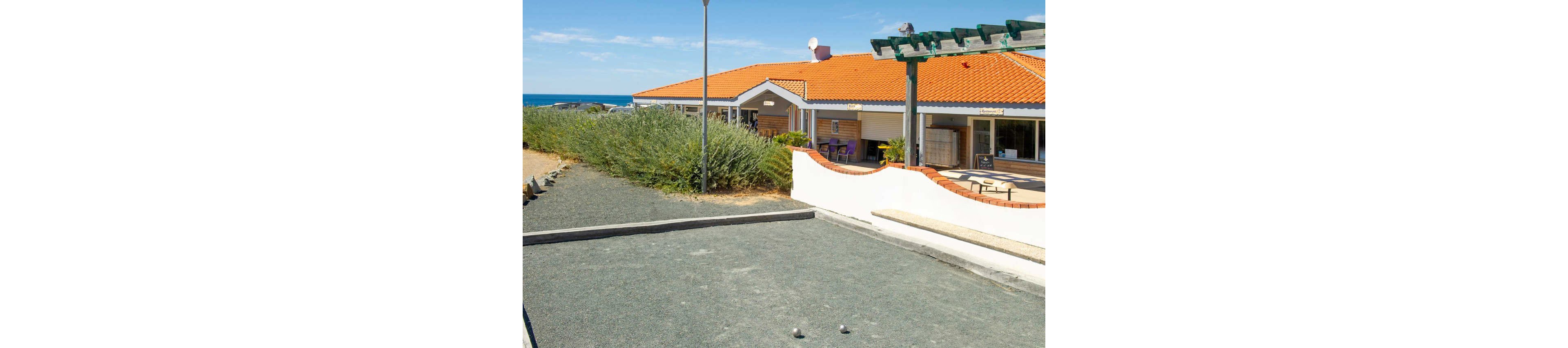 A group of adults and children play pétanque on a gravel court at a seaside holiday resort. The players are casually dressed in summer clothing, with one child preparing to throw a metal ball. Behind them, low buildings with orange-tiled roofs and outdoor seating suggest a relaxed vacation setting near the coast. Bright sunlight, blue sky, and greenery create a cheerful, summery atmosphere.