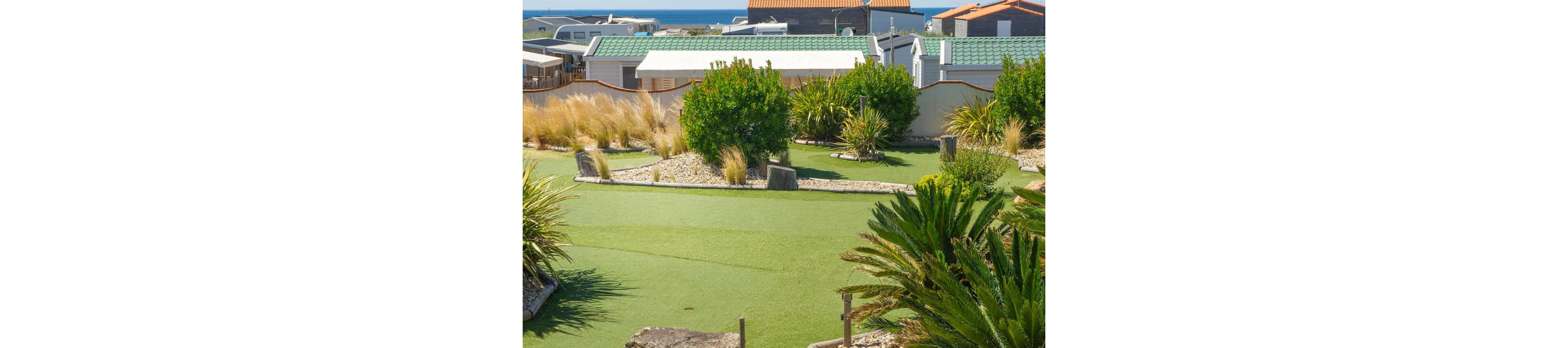 A family group plays a round of mini golf on a sunny day. Children and adults gather around a putting green as one person lines up a shot while another keeps score. The course is surrounded by lush plants, rocks, and ornamental grasses. In the background, holiday cabins and the distant sea suggest a relaxed coastal vacation setting.