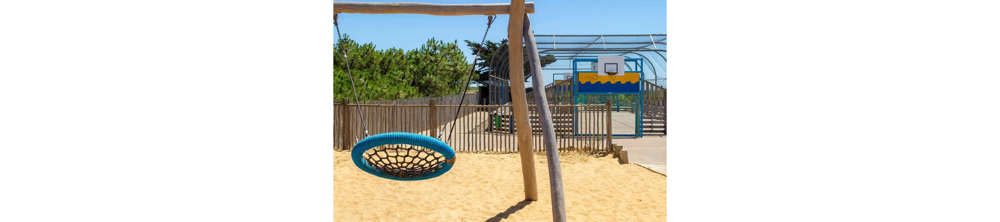 Several children play on a large basket swing in a sunny outdoor playground. A child in a red dress stands in the sand pushing the swing while two others sit and hold onto the ropes. In the background, more children walk near a fenced sports court with a basketball hoop and colorful backboard. The scene is bright and cheerful with clear blue skies and trees surrounding the play area.