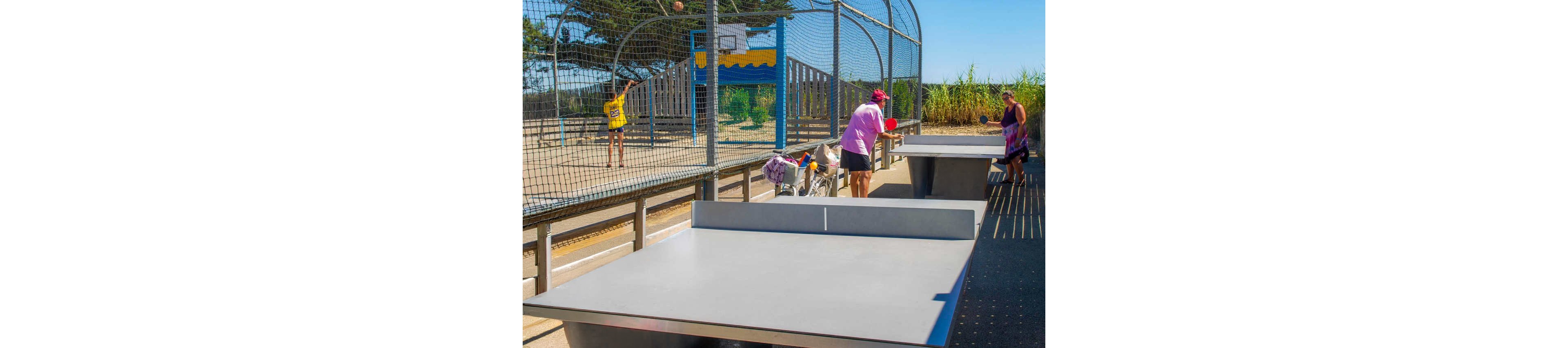 Several people gather around an outdoor table tennis table in a sunny park. Children and adults hold paddles, ready to play or watching the game. A fenced sports court and basketball hoop appear in the background, along with greenery and a bright blue sky. The scene suggests a casual, active day out with family and friends.