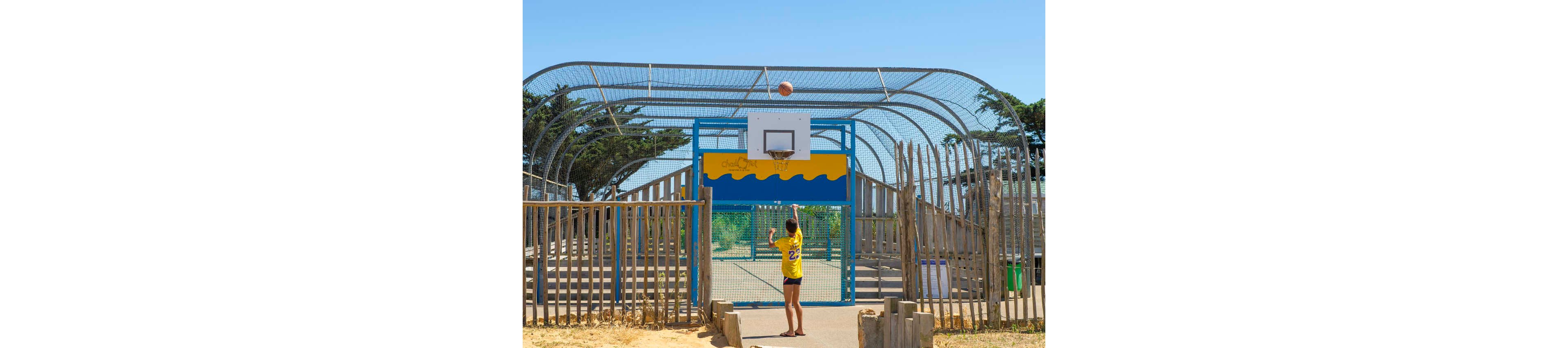 A teenager in a yellow jersey shoots a basketball toward a hoop on an outdoor court. The court is enclosed with metal fencing and surrounded by wooden posts, giving it a beachside or park-like atmosphere. Bright sunlight and clear blue sky create a vibrant, summery feel. The player stands centered beneath the hoop, emphasizing action and recreation in an urban outdoor setting.