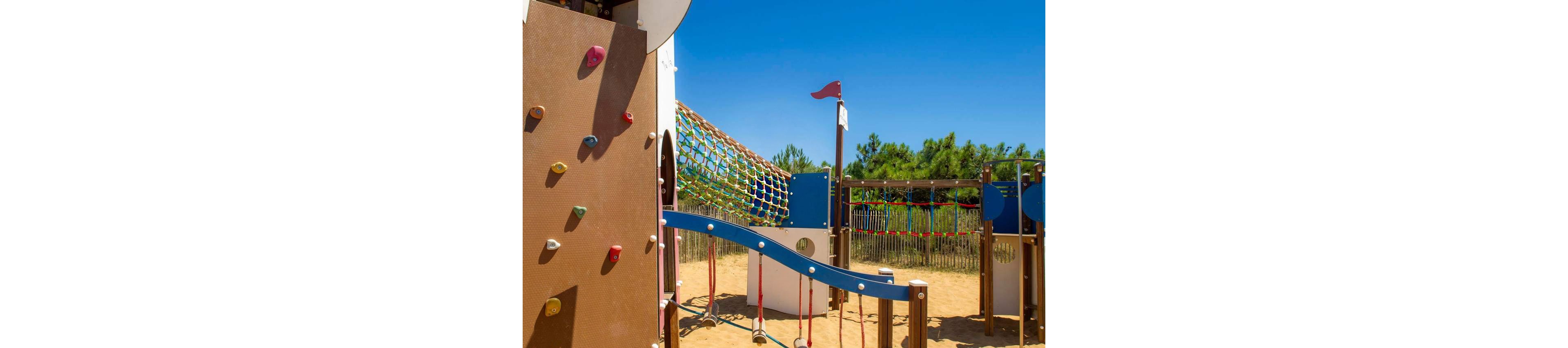 Children are climbing and exploring a colorful outdoor playground structure. The scene shows a sandy play area with climbing walls, rope tunnels, and elevated walkways. Bright summer sunlight and a clear blue sky create a warm, cheerful atmosphere. Trees and a wooden fence in the background suggest the playground is located in a park or recreational area.