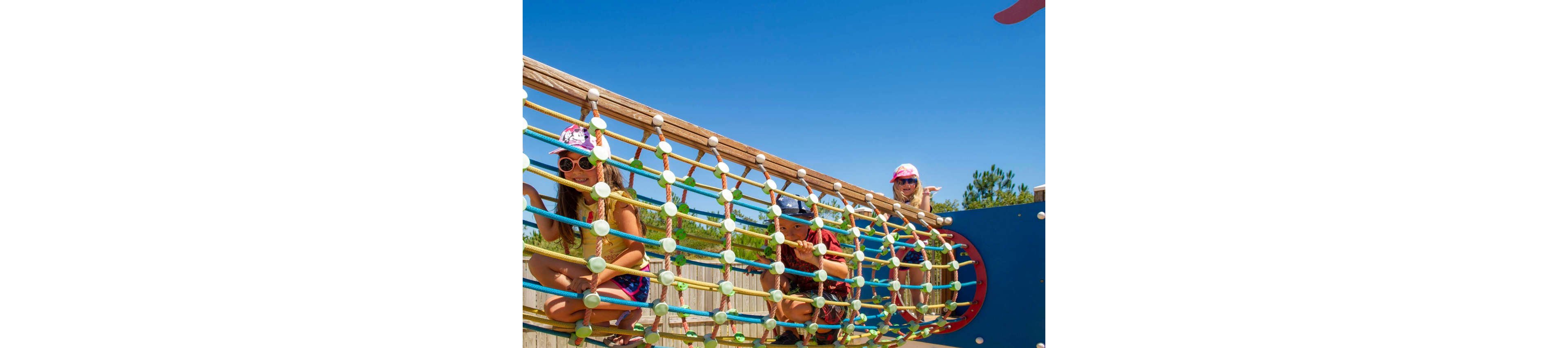 Children play on a colorful rope tunnel structure at an outdoor playground. They crawl and climb through the netting under a bright blue sky. The play equipment features multicolored ropes and a large circular blue panel. A wooden fence and trees are visible in the background, emphasizing the sunny, outdoor setting.