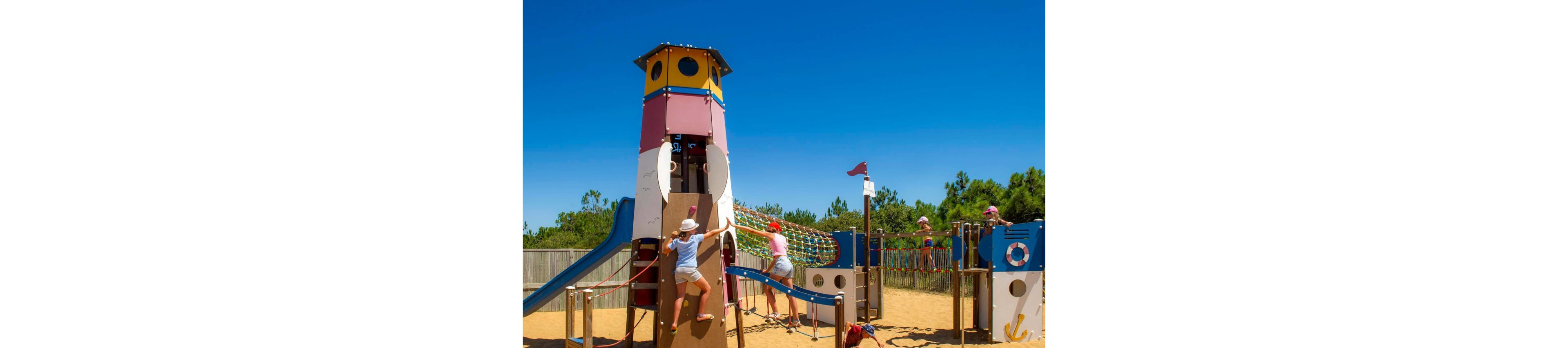 Several children play on a large, colorful playground structure designed like a ship or lighthouse in a sandy outdoor area. The kids climb ropes, ladders, and platforms under a bright blue sky. The playground is surrounded by a wooden fence and trees in the background. The scene feels energetic, sunny, and summery, with children wearing hats and casual clothes for outdoor play.