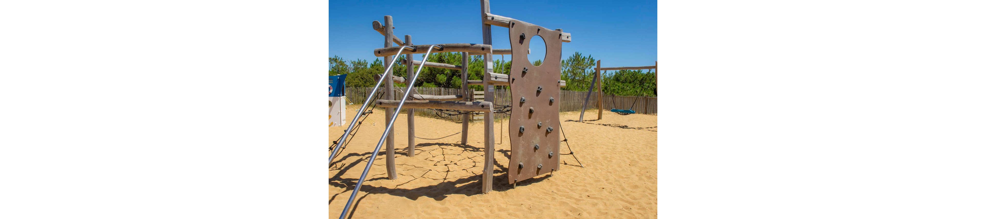 Several children play on a wooden playground structure in a sandy outdoor area. Two kids climb a vertical climbing wall while another hangs upside down from a metal bar. The scene is set under a clear blue sky with trees and a wooden fence in the background. Bright summer clothing and strong sunlight create a cheerful, energetic atmosphere.