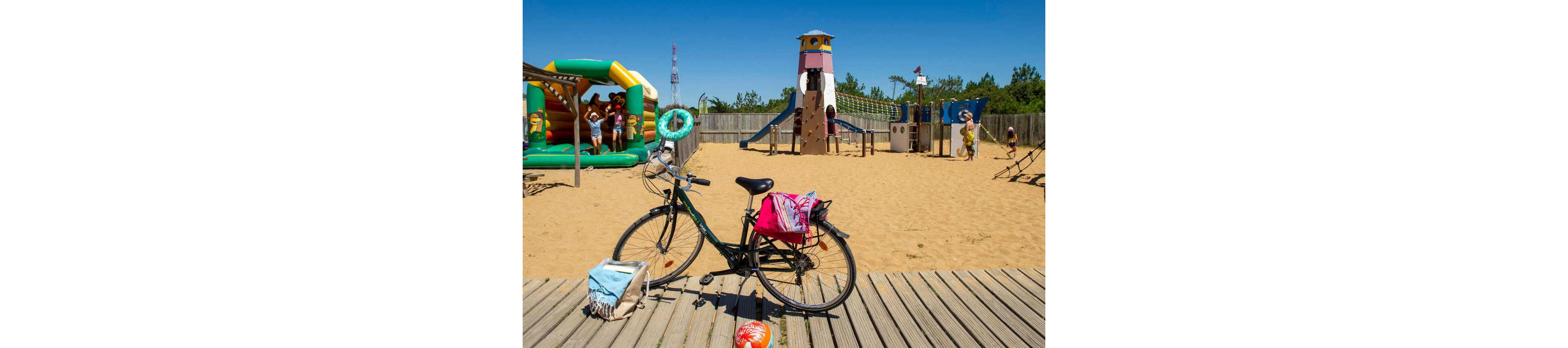 A bicycle with bags and a towel is parked on a wooden boardwalk beside a sandy playground. In the background, children play on a colorful inflatable bounce house and a tall slide structure under a clear blue sky. The fenced area includes various play equipment and scattered chairs, suggesting a family-friendly recreational spot. Bright sunlight and vivid colors create a cheerful summer atmosphere.