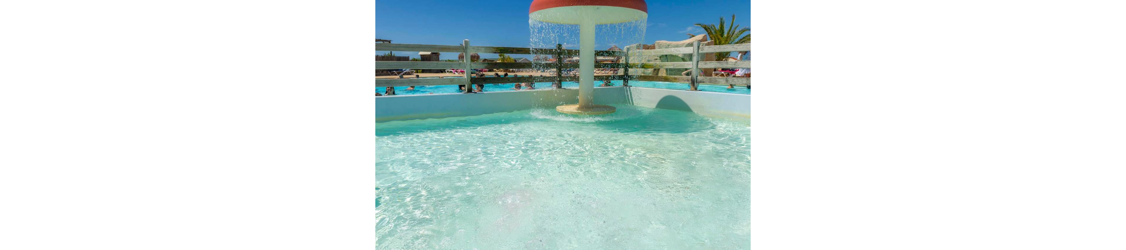 Two young girls play together in a shallow splash pool under a mushroom-shaped water feature. An adult relaxes nearby in the water while other visitors enjoy a larger pool in the background. The scene is outdoors on a sunny day with clear blue sky and palm trees, suggesting a warm vacation setting. Bright colors and sparkling water create a cheerful, family-friendly atmosphere.