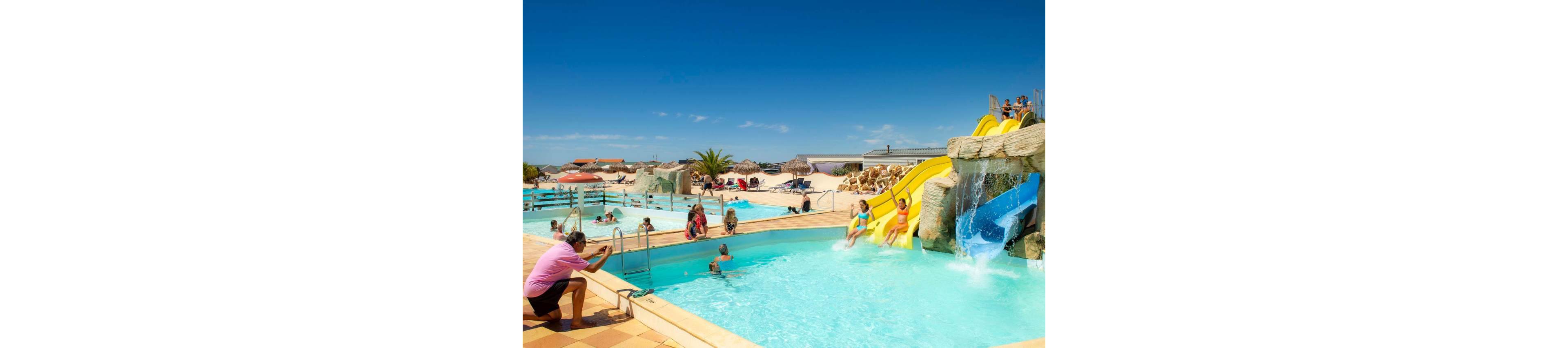 Families and children play in a bright outdoor water park with multiple swimming pools. Several kids slide down yellow water slides built into artificial rock formations, splashing into the clear blue water. Adults relax in the pool and on the surrounding deck, while one person takes photos near the edge. In the background, sun loungers, umbrellas, and sandy areas create a beach resort atmosphere under a clear blue sky.