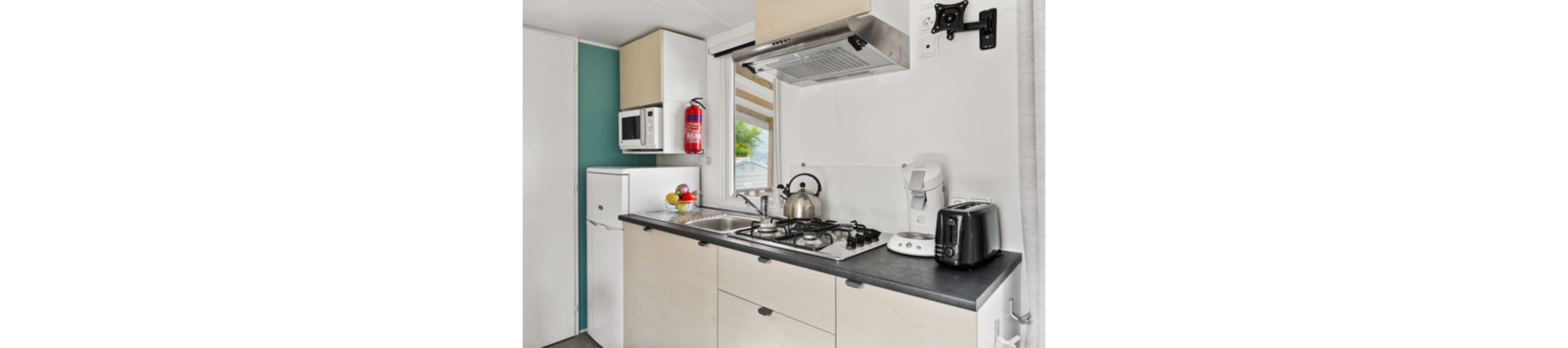 A compact modern kitchenette features light wood cabinets and a dark countertop. The space includes a gas cooktop with a stainless steel kettle, a toaster, and a coffee maker. A small refrigerator, microwave, and wall-mounted range hood complete the functional setup. A bowl of fresh fruit and a window with striped blinds add color and natural light to the clean, minimalist interior.