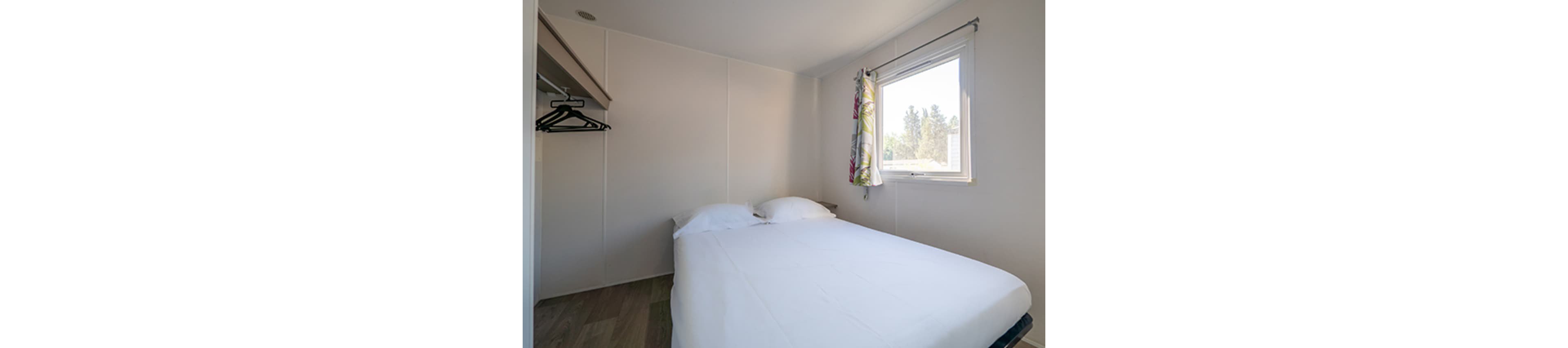 A compact bedroom features a neatly made double bed with white linens against plain light-colored walls. Natural light enters through a single window with a floral curtain on one side. An open wardrobe with several black clothes hangers is visible to the left. The room has a simple, minimalist design with a calm and uncluttered atmosphere.