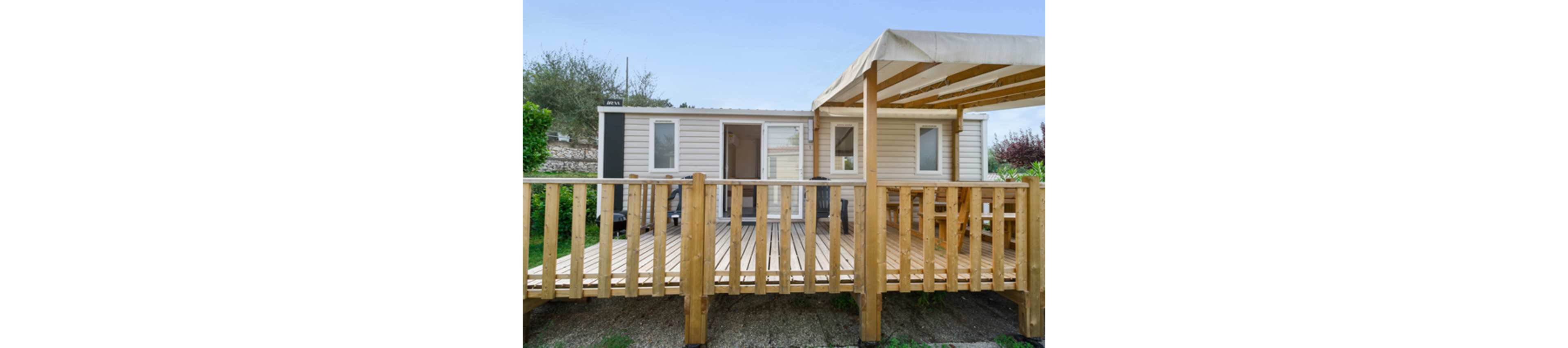 A light-colored mobile home features a spacious wooden deck in the foreground. The terrace is enclosed by a simple wooden railing and partially covered by a pergola-style canopy. The structure sits on a gravel base with greenery and trees visible in the background. The scene suggests a quiet vacation setting or residential campsite under a clear sky.