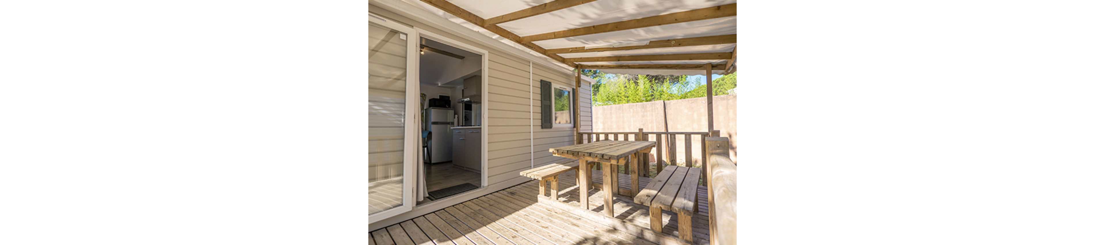 A wooden terrace features a sturdy picnic-style table and benches under a partial pergola roof. The space is attached to a light-colored mobile home or cabin with sliding glass doors leading to the interior. Inside, a compact kitchen area with appliances is visible. Bright sunlight and greenery beyond the fence create a relaxed, vacation-like atmosphere.