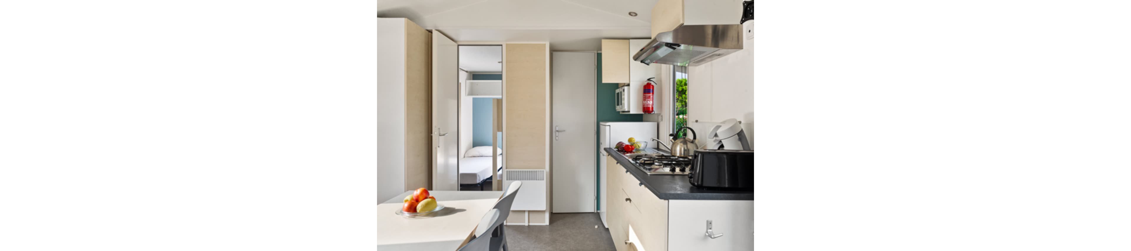 Bright, compact kitchen interior in a modern mobile home or small apartment. The space features a gas cooktop with kettle, microwave, refrigerator, and storage cabinets. A dining table with chairs and a bowl of fresh fruit sits in the foreground. A bedroom with a neatly made bed is visible through an open doorway, and a red fire extinguisher hangs on the wall near the kitchen.