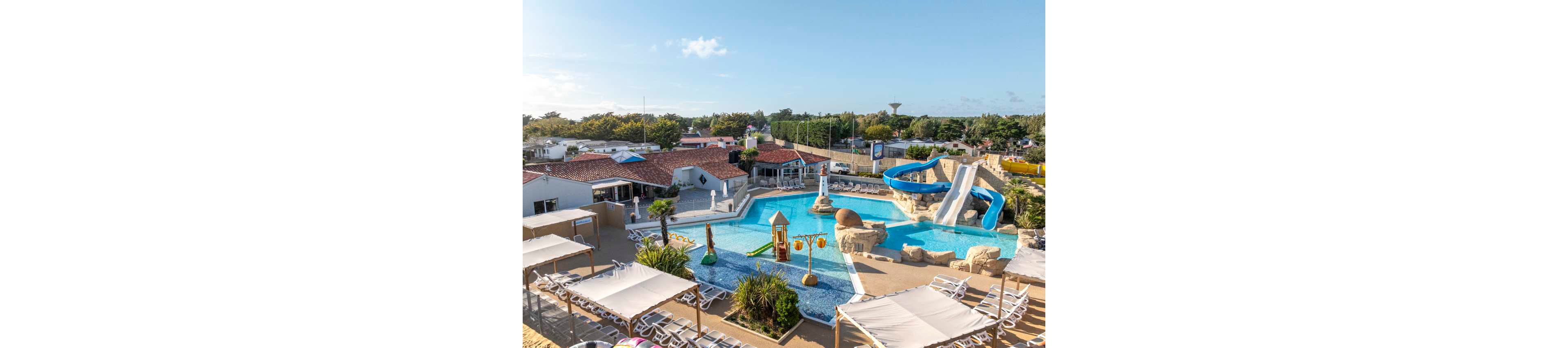 An outdoor resort swimming pool features winding blue waterslides and a shallow play area with fountains and water toys. Rows of sun loungers and shaded canopies surround the pool, creating a relaxing space for guests. Low-rise buildings with red-tiled roofs and lush greenery frame the scene in the background. The bright daylight and clear sky give the setting a warm, inviting holiday atmosphere.