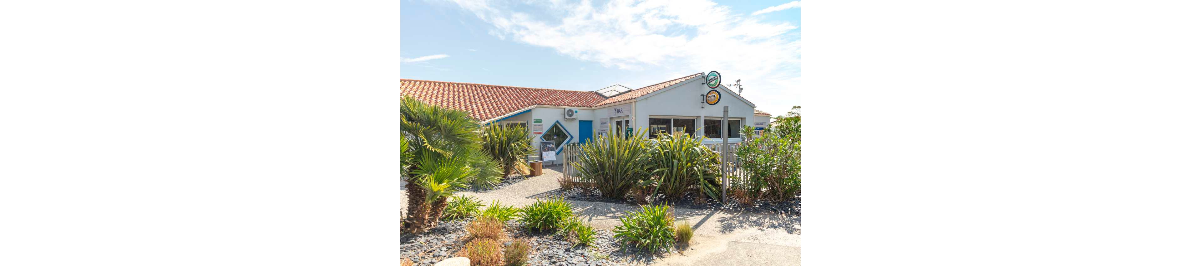 A small white café building with a red-tiled roof sits under a bright blue sky. The entrance is surrounded by dense green plants and palm-like shrubs, giving a coastal, Mediterranean feel. Outdoor signage and a menu board are visible near the doorway, suggesting a casual dining or refreshment spot. The overall atmosphere is relaxed and inviting, ideal for vacation or seaside leisure.