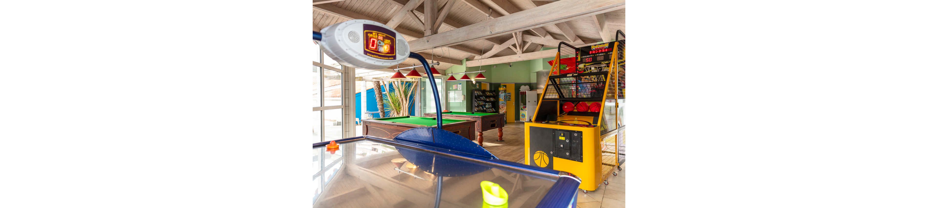 An indoor arcade room features an air hockey table in the foreground and a basketball arcade game to the right. A green pool table and additional game machines are visible toward the back of the room. Large windows let in natural light, illuminating wooden ceiling beams and tiled floors. Red hanging lamps and colorful machines create a playful, family-friendly atmosphere.
