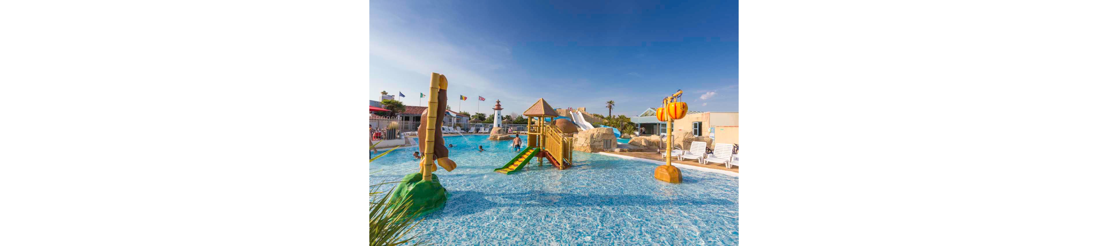 An outdoor family water park features a shallow pool with children playing around colorful slides and climbing structures. The scene includes a small lighthouse, rock formations, and multiple water slides in the background. International flags line the top of nearby buildings under a clear blue sky. Lounge chairs and umbrellas surround the pool area, suggesting a relaxed vacation atmosphere.
